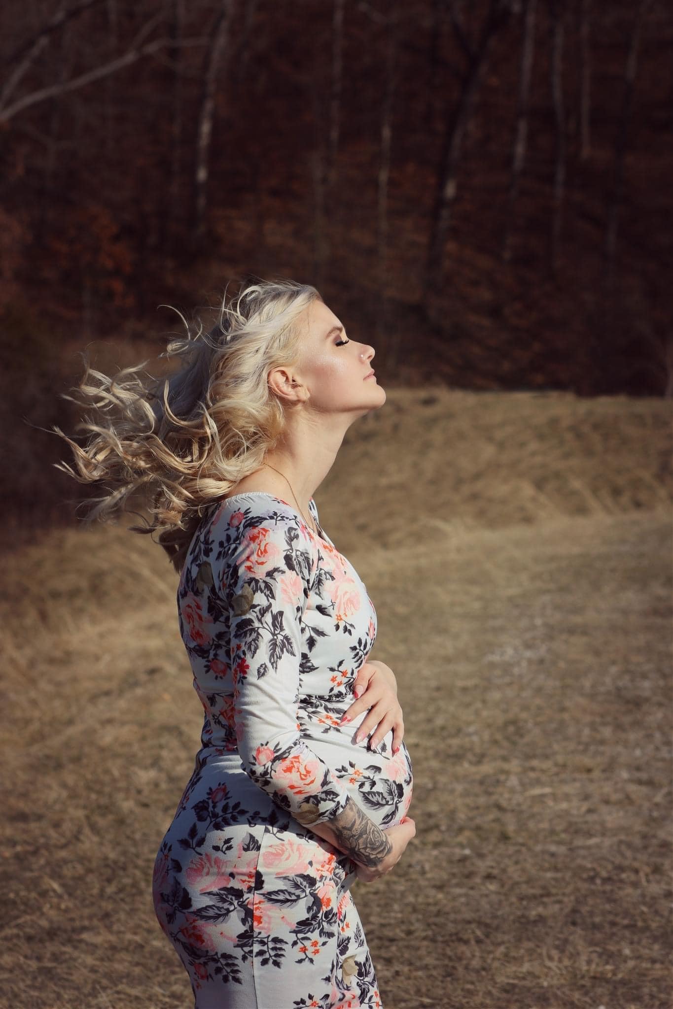 Expectant mother in a floral dress, eyes closed, hair blowing in an open field